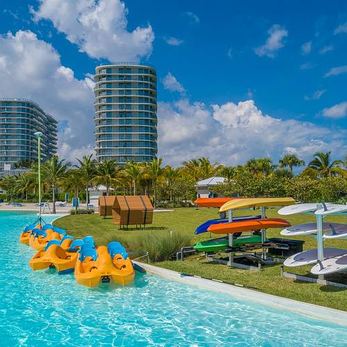 Shopping. Dining. Gyms. Walking Trails. Playgrounds. Paddle. It's Your Perfect Day at SoLe Mia. A sunny poolside scene with kayaks, paddleboards, and modern buildings in the background, surrounded by lush greenery.
